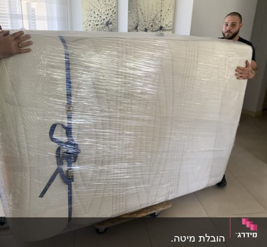 שני אנשים מעבירים מזרן עטוף בניילון על עגלה